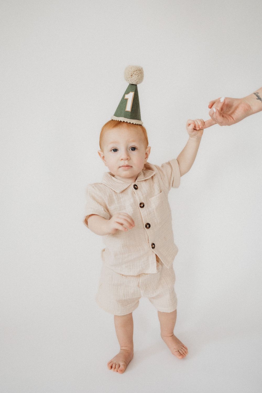 A baby in a beige linen outfit wearing a green number 1 birthday party hat celebrates their first birthday.