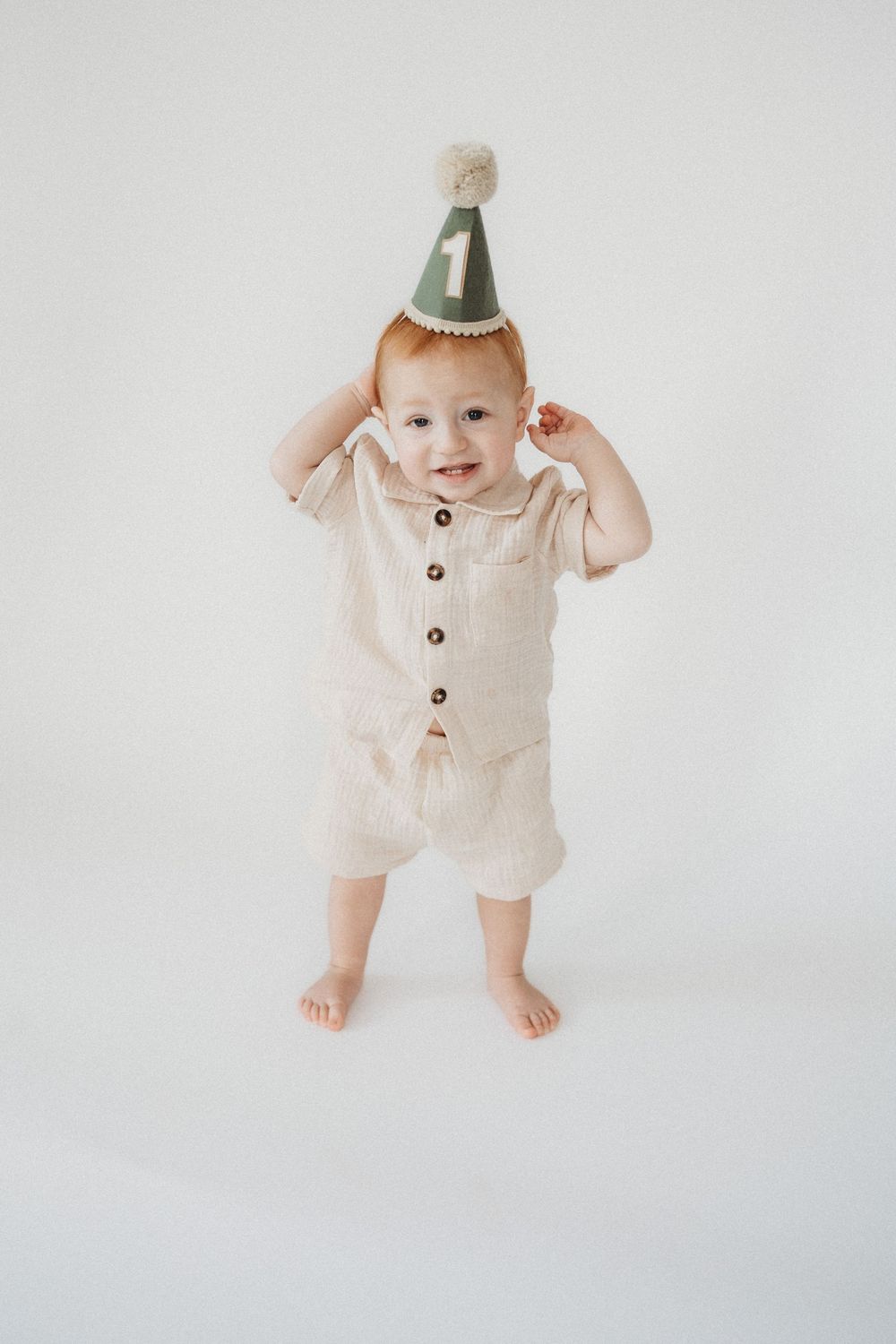 Smiling baby in beige linen outfit and green number one birthday party hat standing barefoot on white background.
