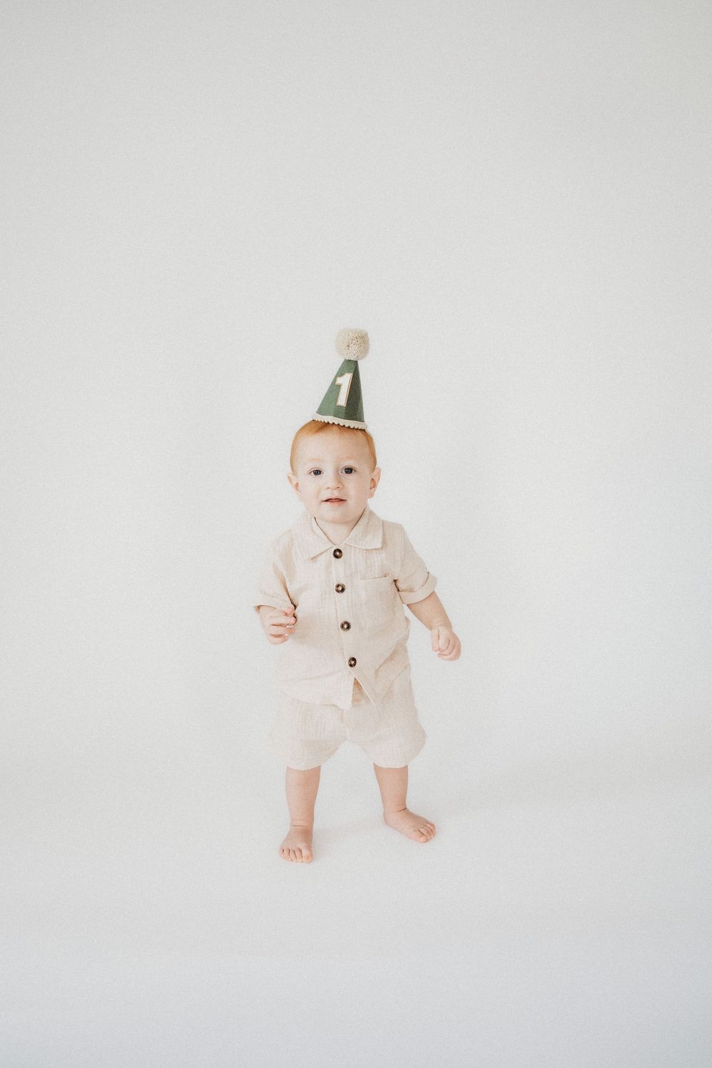 A baby in a beige romper wearing a green party hat with the number 1, standing on a white background.
