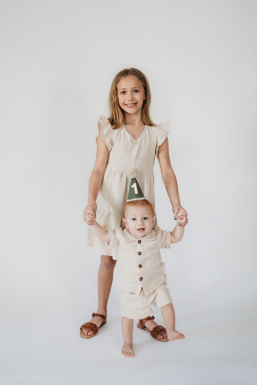 A smiling girl in a beige dress helps a baby in a linen outfit and birthday crown stand on a white background.