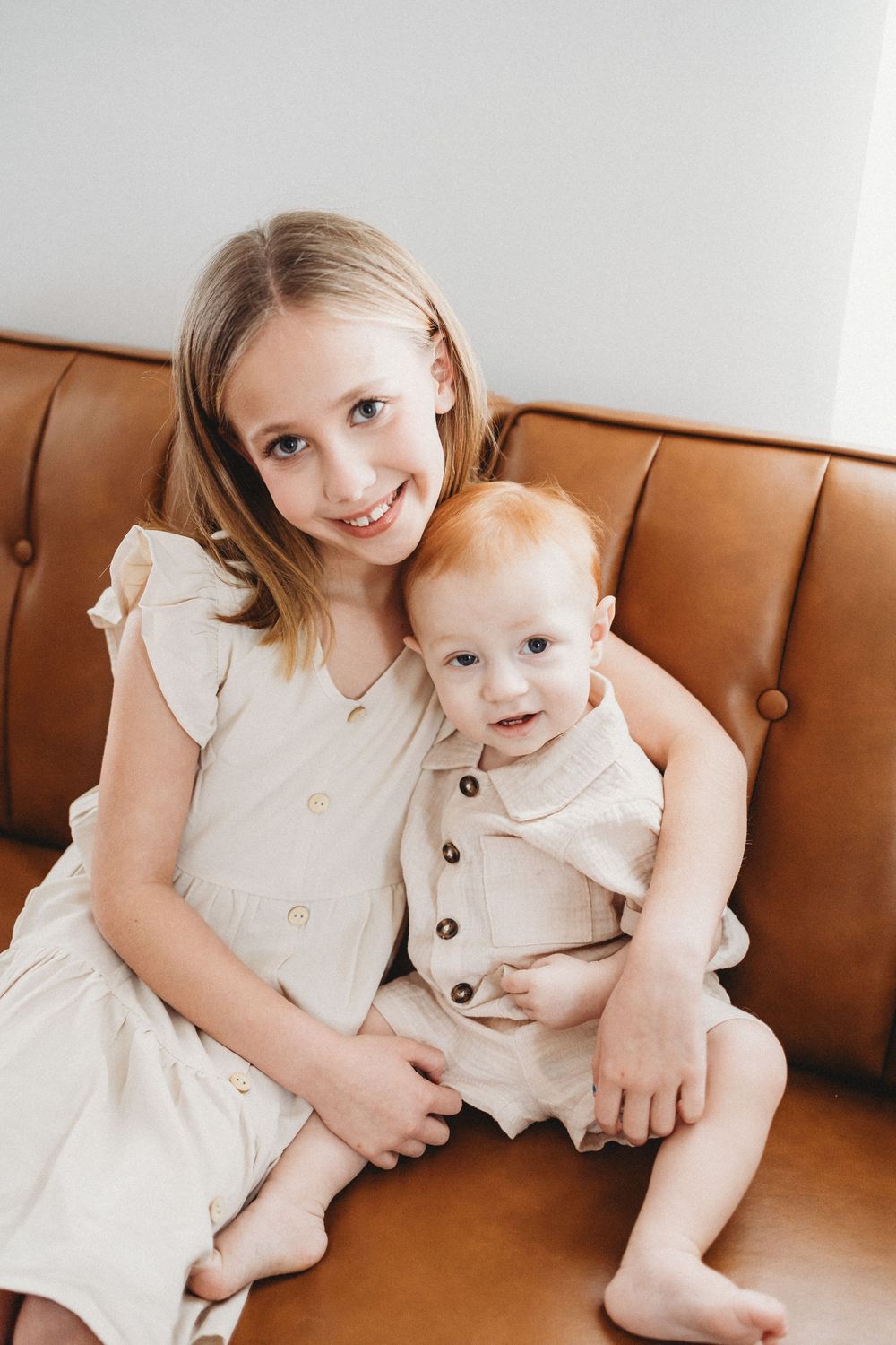 Smiling girl in cream dress hugging a baby on a brown leather sofa.