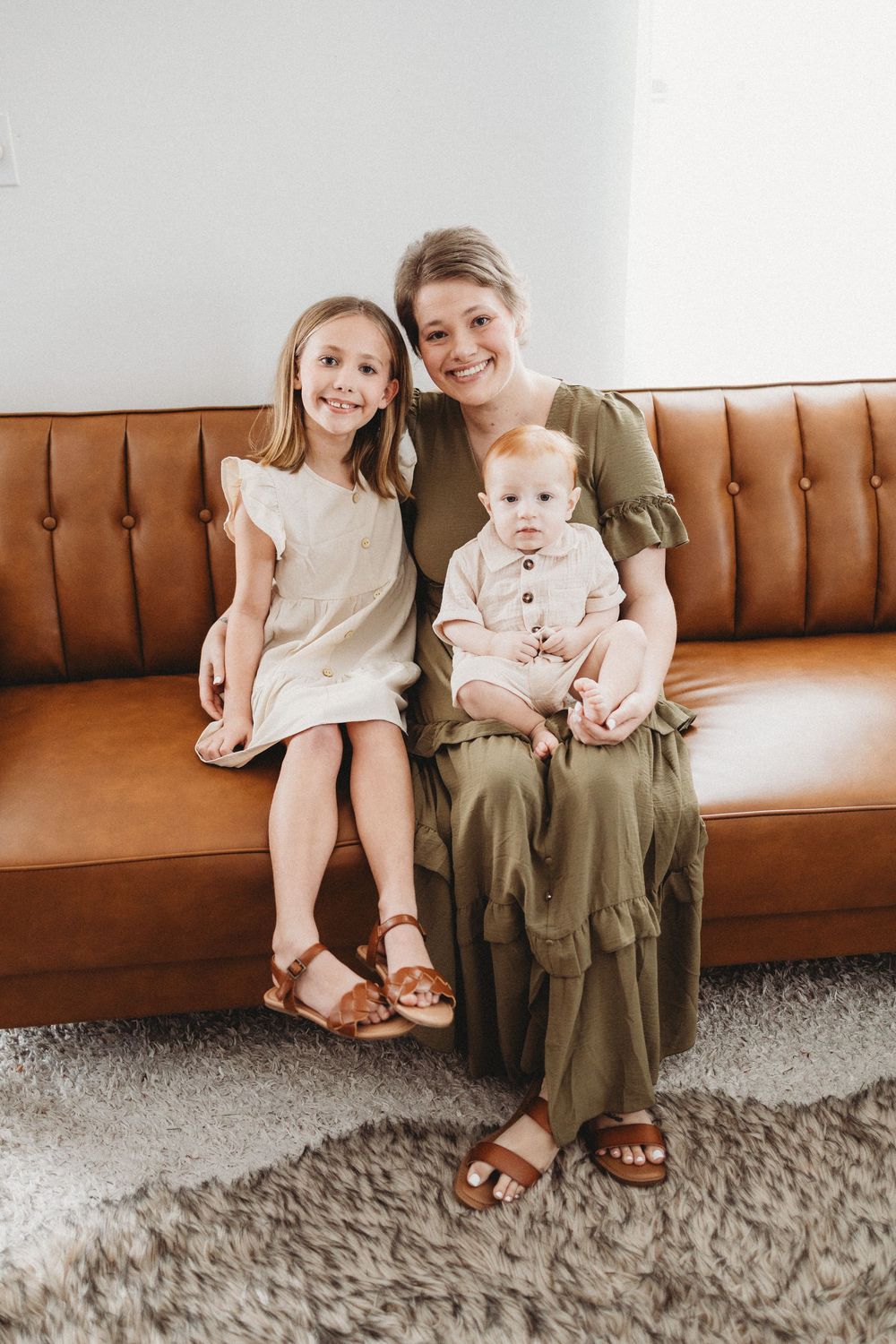 A smiling woman in olive dress sits on brown leather sofa holding baby, with young blonde girl in white dress beside her.