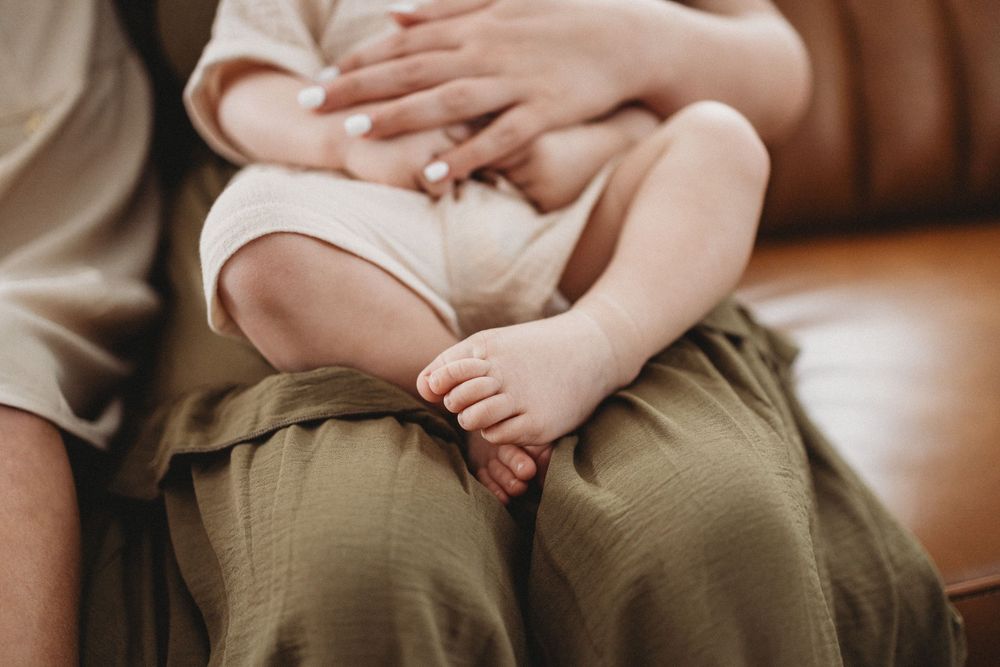 Close-up of a baby sitting on a parent's lap in an olive green skirt, with tiny feet and hands visible.