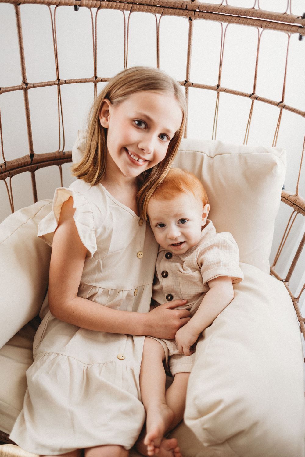 A smiling girl in a white dress holds a red-haired baby in a wicker chair, both wearing neutral tones.