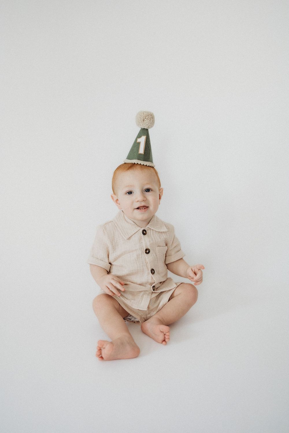 A baby wearing a green number 1 birthday hat and beige romper sits on a white background.