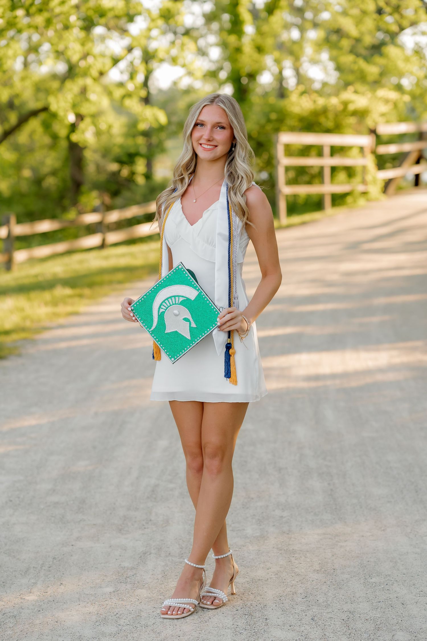 Smiling blonde graduate in white dress holds Michigan State cap and tassel on a tree-lined gravel path.