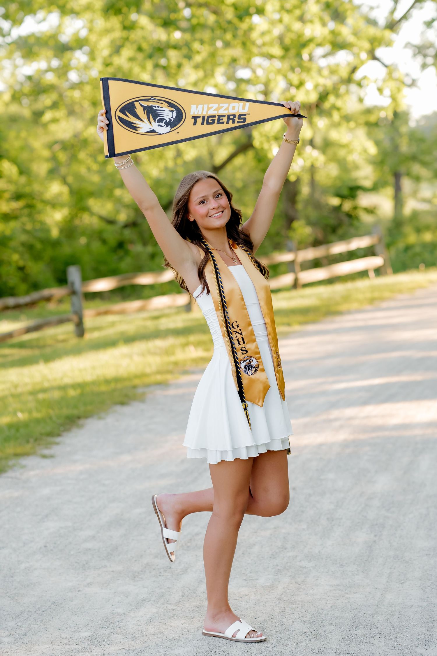 Young woman in white dress holding Mizzou Tigers pennant, wearing graduation sash, celebrating outdoors on a tree-lined path.