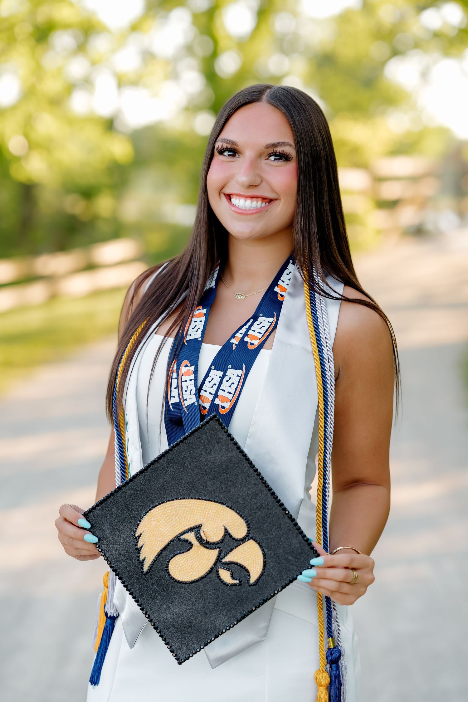Smiling graduate holds University of Iowa Hawkeyes mortarboard cap wearing white gown and blue gold honor cords outdoors.