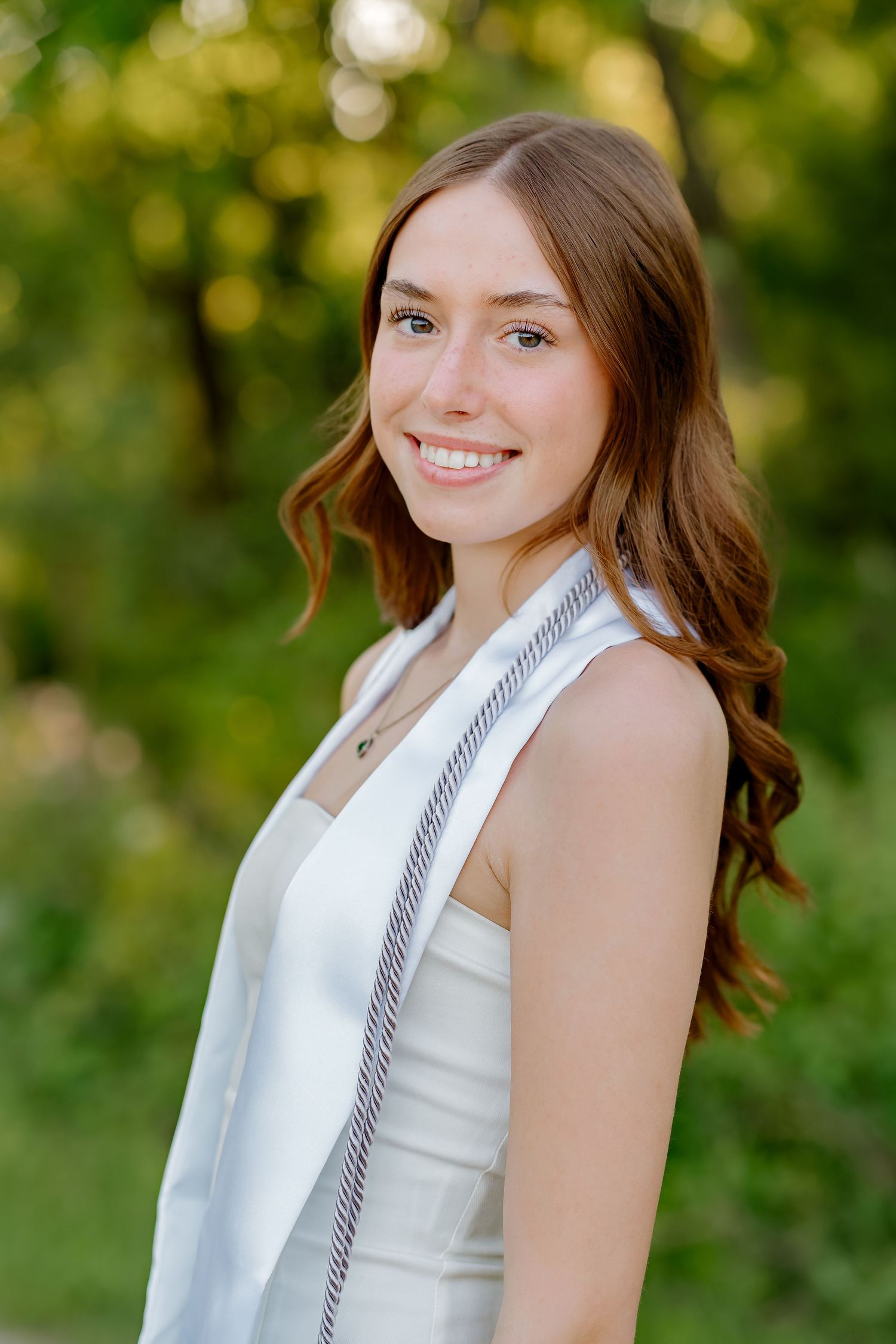 Smiling young woman in white dress and graduation sash poses outdoors with green trees in background.