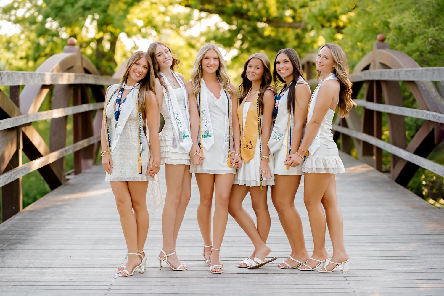 Six young women in white dresses and graduation sashes pose together on a wooden bridge outdoors.