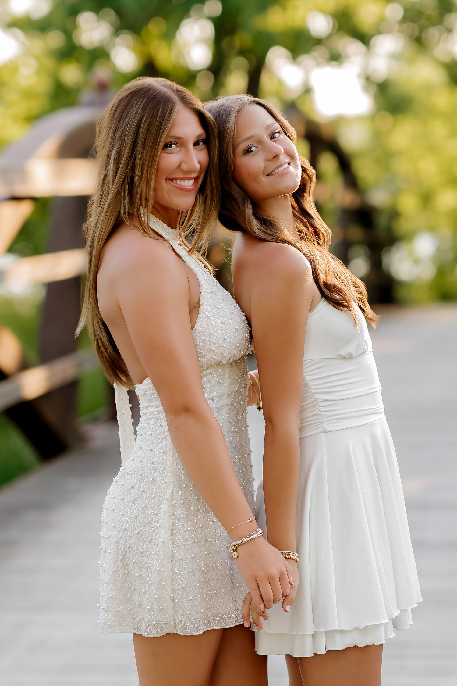 Two young women in white dresses smiling outdoors on a bridge surrounded by green trees.