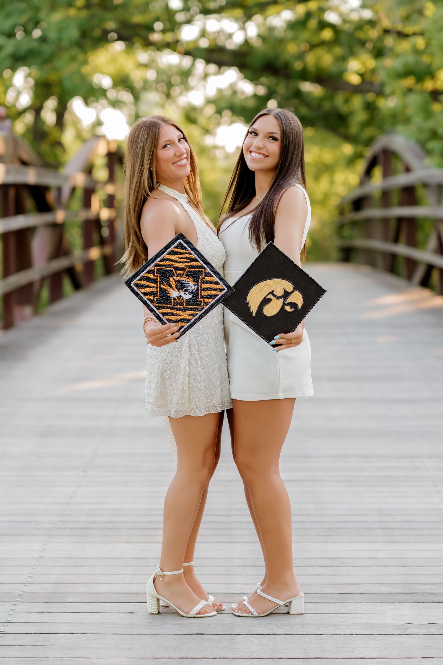 Two smiling graduates pose on a wooden bridge holding decorated caps from Missouri and Iowa universities.