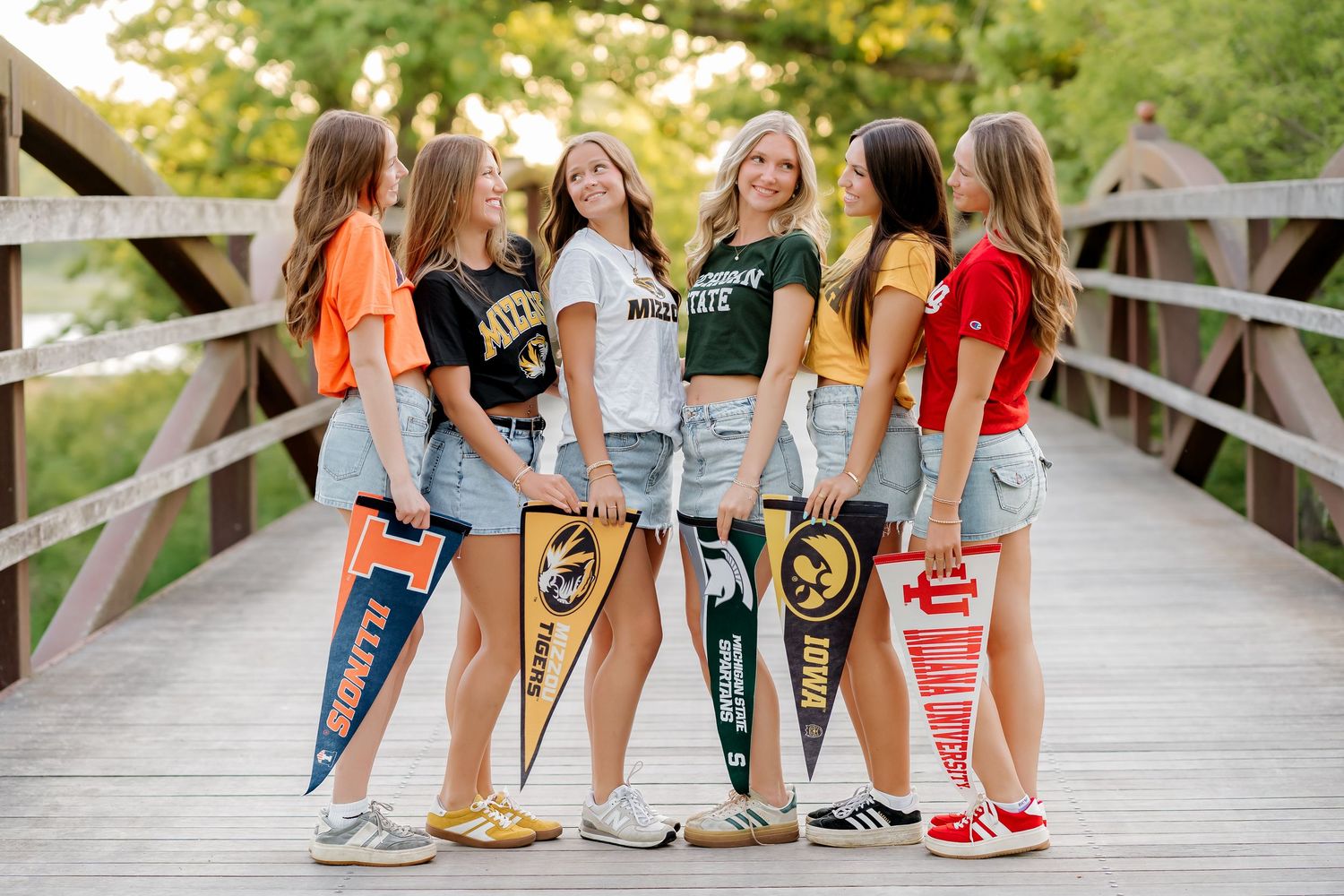 Six young women pose on a wooden bridge holding college pennants, wearing casual summer outfits with denim shorts.
