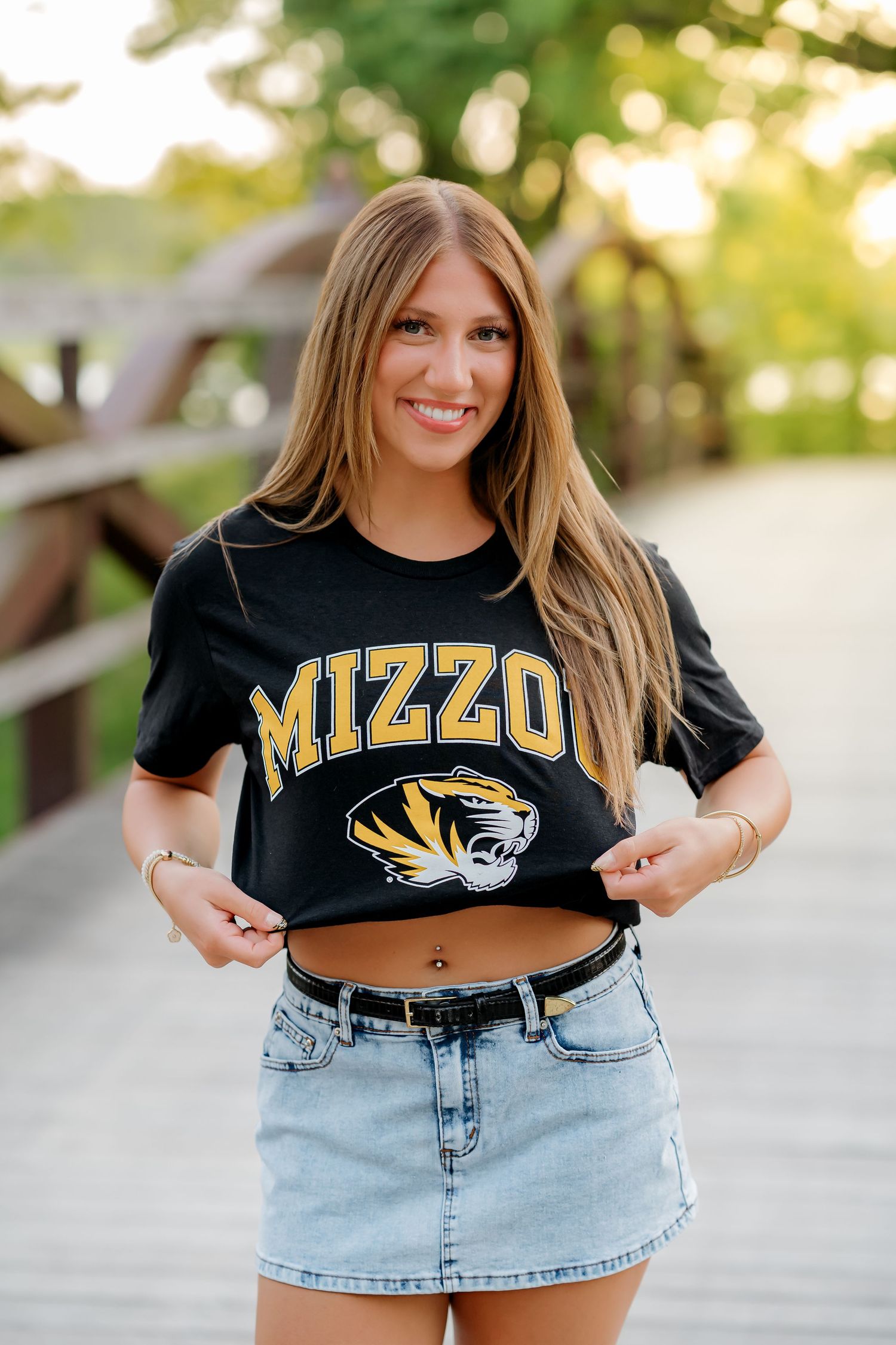 Smiling young woman on a bridge wearing a black Mizzou Tigers crop t-shirt with a denim skirt and belt.