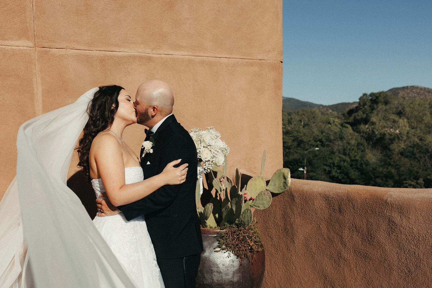Bride and groom share a romantic kiss on a terrace with mountain views and adobe wall backdrop on their wedding day.