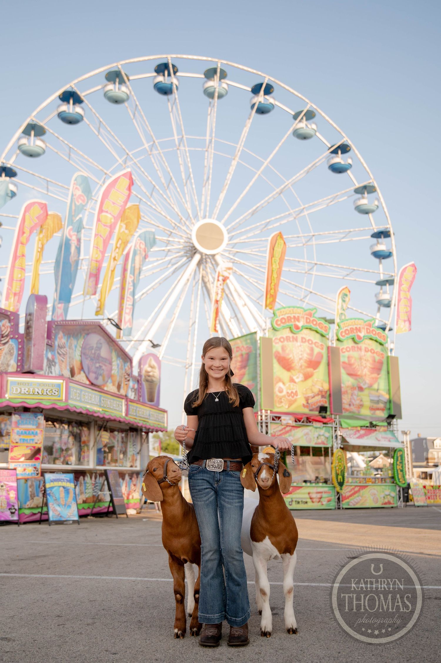 Young girl holds two goats at a county fair with a large Ferris wheel and colorful carnival games in the background.
