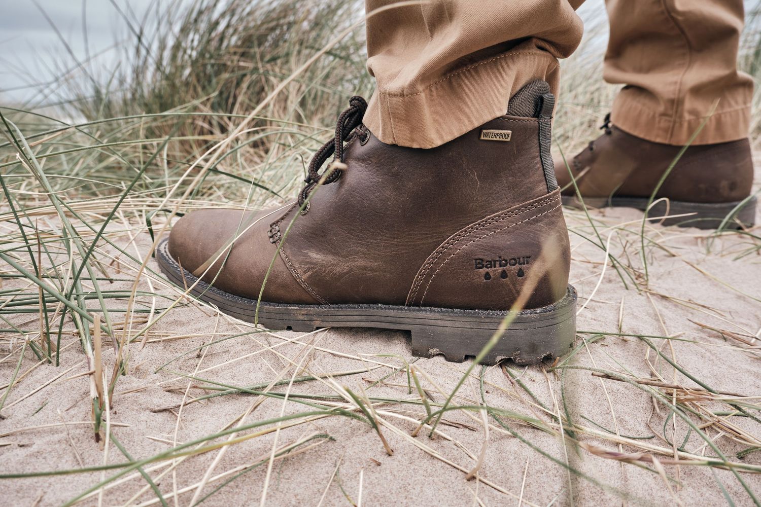 Brown Barbour lace-up leather boots worn on sandy beach with grass, paired with tan trousers.