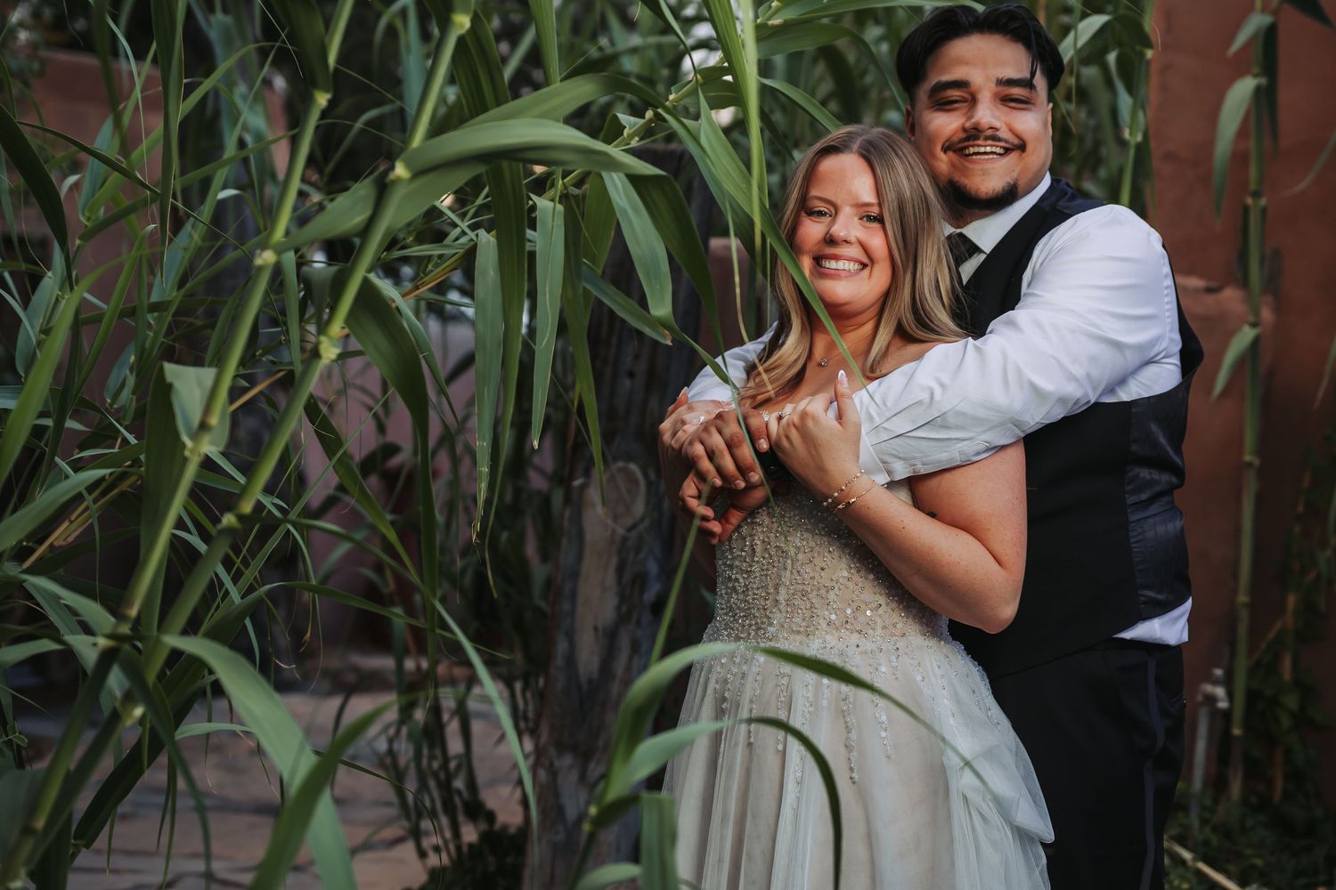 A smiling bride in a white gown embraces her groom in a vest among lush green bamboo plants.