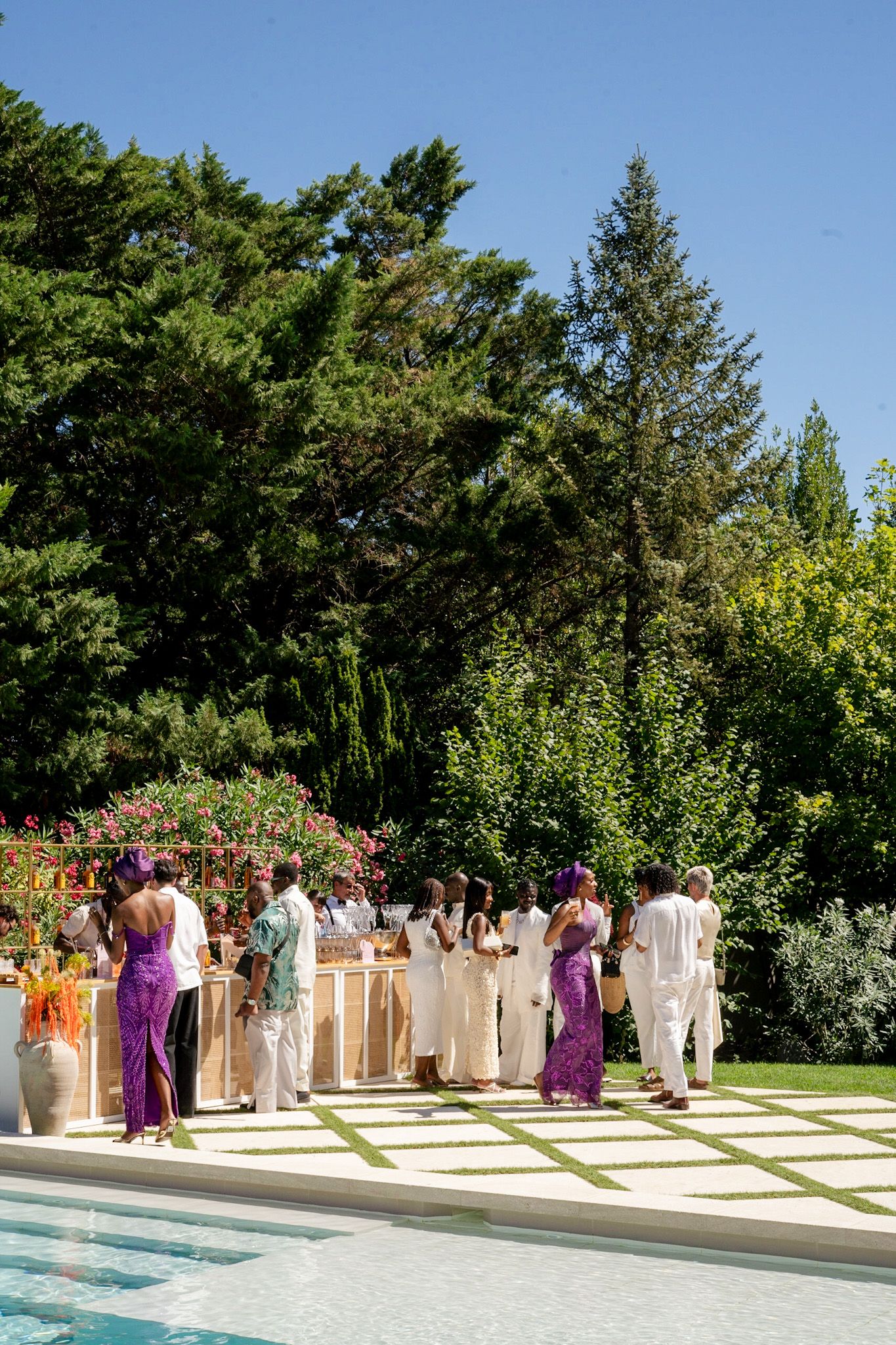 Elegant outdoor poolside gathering with guests in white and purple attire, surrounded by lush green trees and colorful flowers.