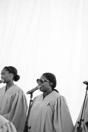 Black and white photo of women in choir robes singing at microphones during an outdoor performance.