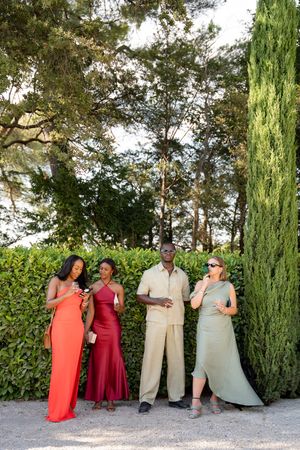 Four elegantly dressed friends socializing outdoors in a lush garden setting, wearing formal attire in red, burgundy, and sage green.