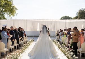 Bride in white gown and veil walks down floral aisle at outdoor wedding ceremony with seated guests.