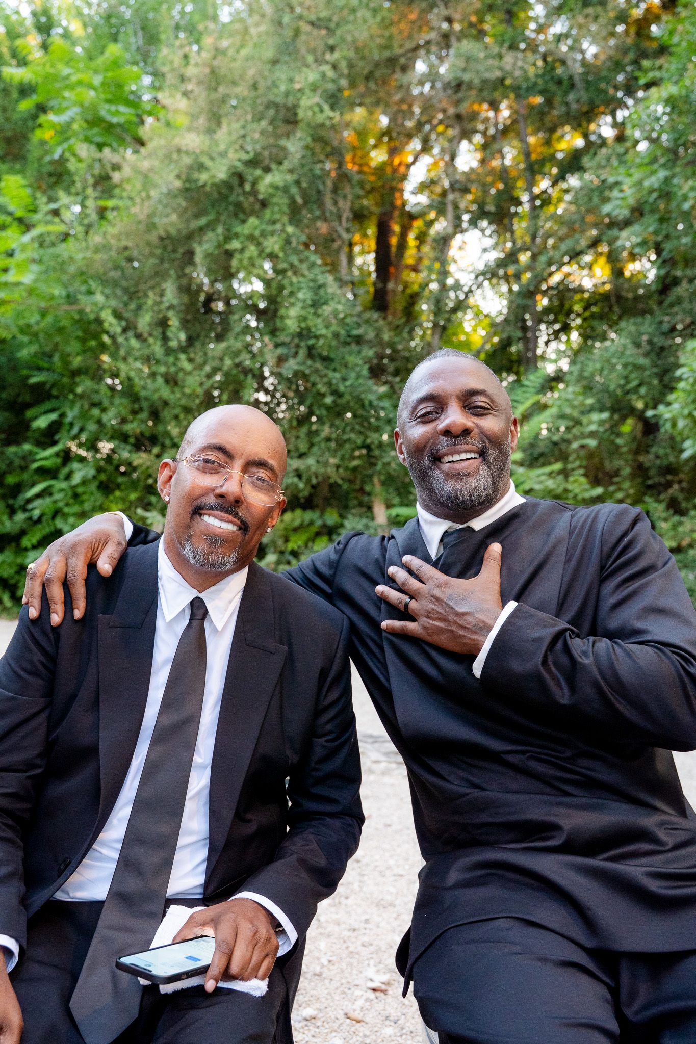 Two well-dressed men in black suits smiling outdoors, posing together with lush green trees in the background.