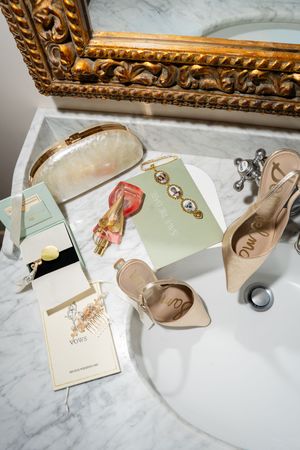 Elegant bridal flat lay on marble sink with nude heels, pearl clutch, perfume, jewelry, and vow booklets.