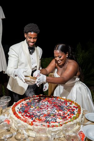 Bride and groom cutting a large fruit-topped wedding cake outdoors at night, both smiling joyfully.