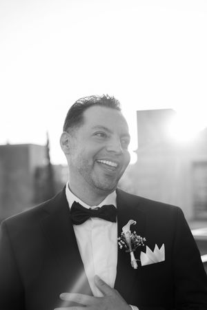 Black and white photo of a smiling groom in a tuxedo with boutonniere and bow tie, posing outdoors with city skyline.