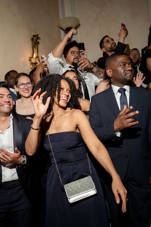 Guests in formal attire celebrate joyfully at an elegant indoor event, with a woman in a navy dress smiling and waving.