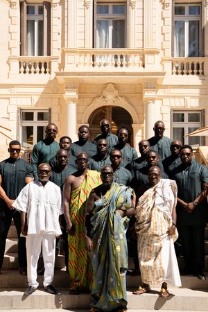 Group of men in traditional African attire and dark outfits posing in front of an elegant building.
