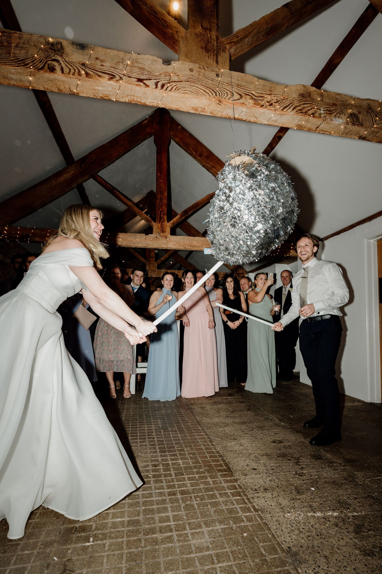 Groom smashing a pinata at a wedding reception in the barn at Quantock Lakes, with guests watching in colorful bridesmaid dresses.