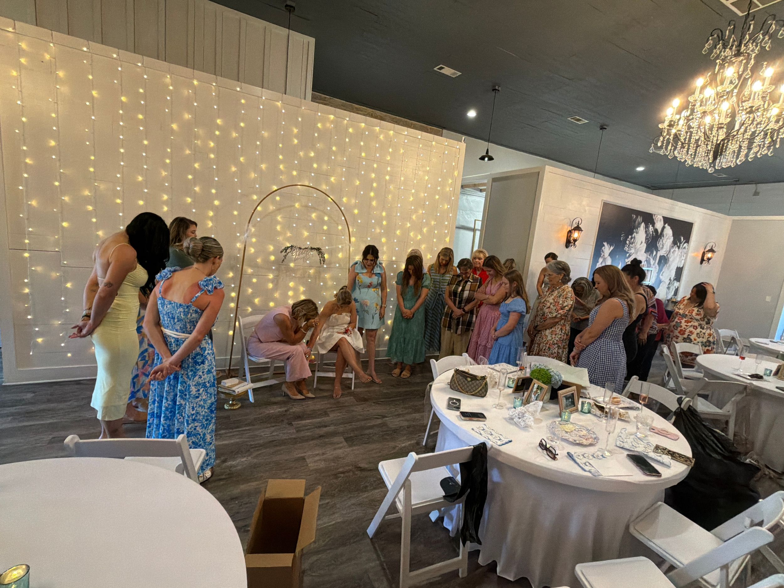 Guests gathered at an elegant baby shower venue with twinkling fairy light backdrop and chandelier lighting.
