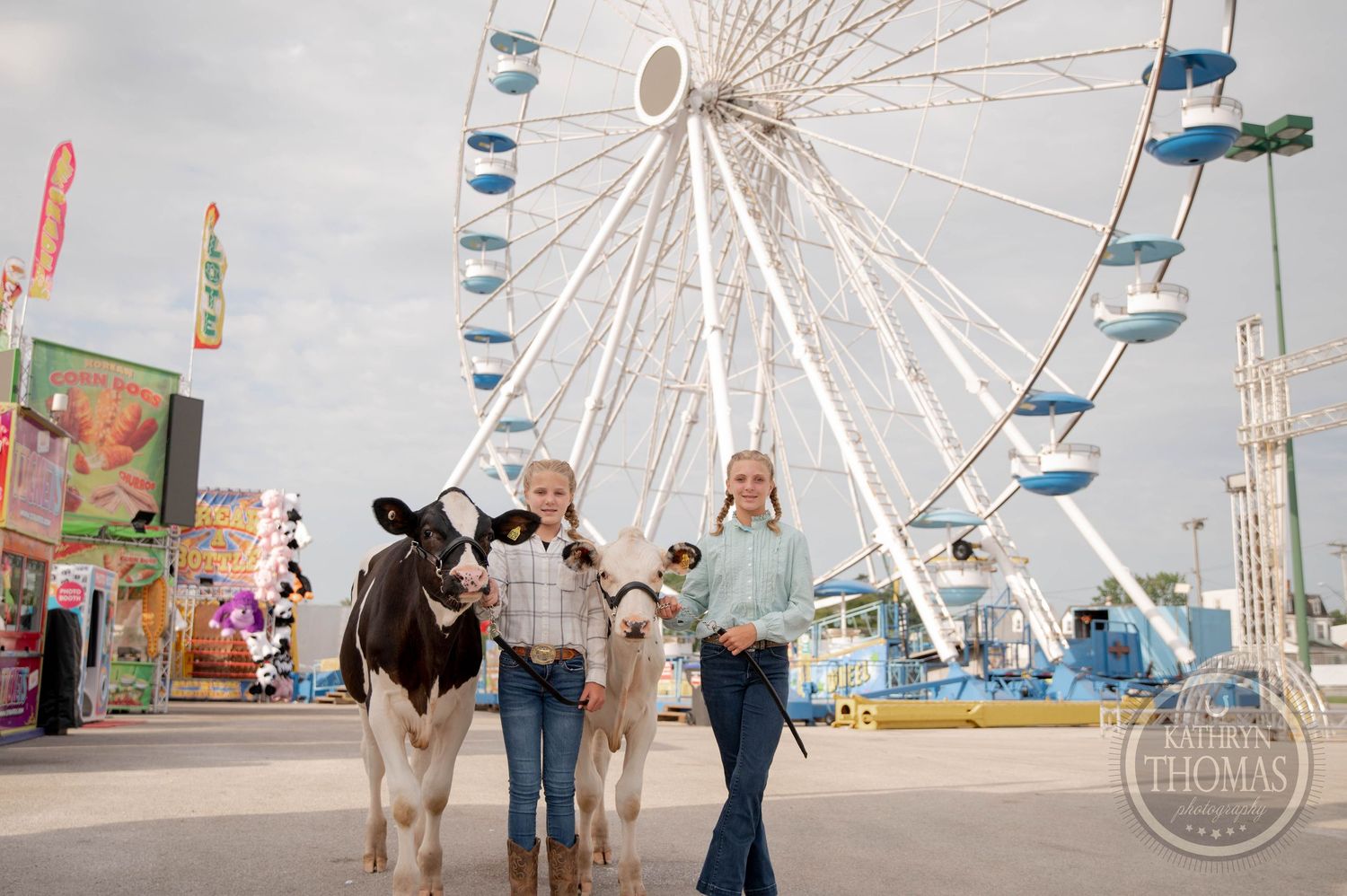 Two people lead a cow and calf under a Welcome to the Fair sign at a colorful county fairground.