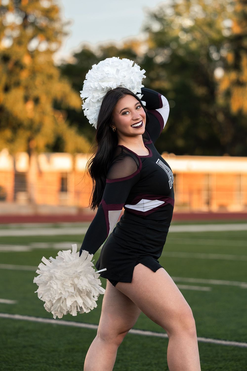 Cheerleader in black uniform performs energetic dance routine with white pom poms on football field at sunset.