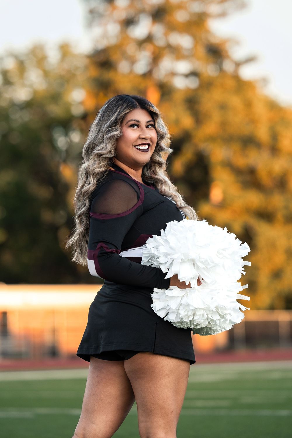 A cheerleader performs energetic dance moves with white pom poms on a sports field during golden hour.