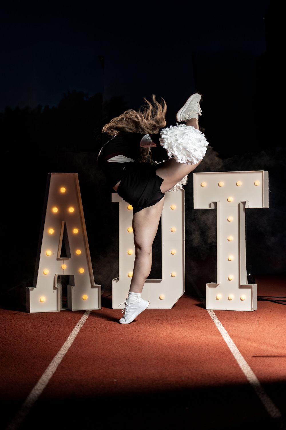 A cheerleader in black uniform performs a high kick pose in front of illuminated marquee letters on a track at night.