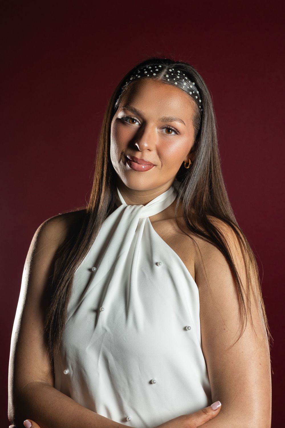 Fashion portrait of a model wearing a white halter top and sparkly headband against a dark burgundy background.