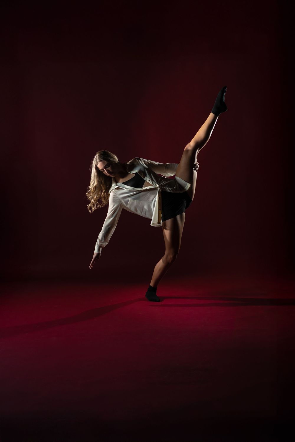Dancer in white top and black shorts performs a dramatic high kick against dark red backdrop.