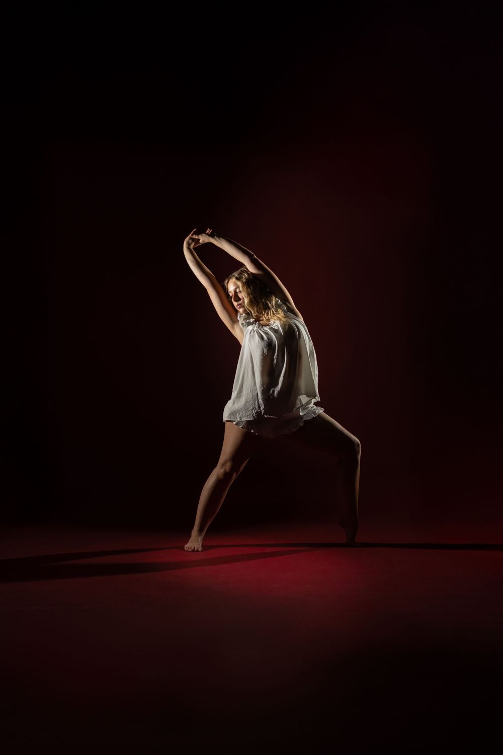 Graceful dancer in white dress performs elegant movement on dark stage with dramatic lighting and red glow.