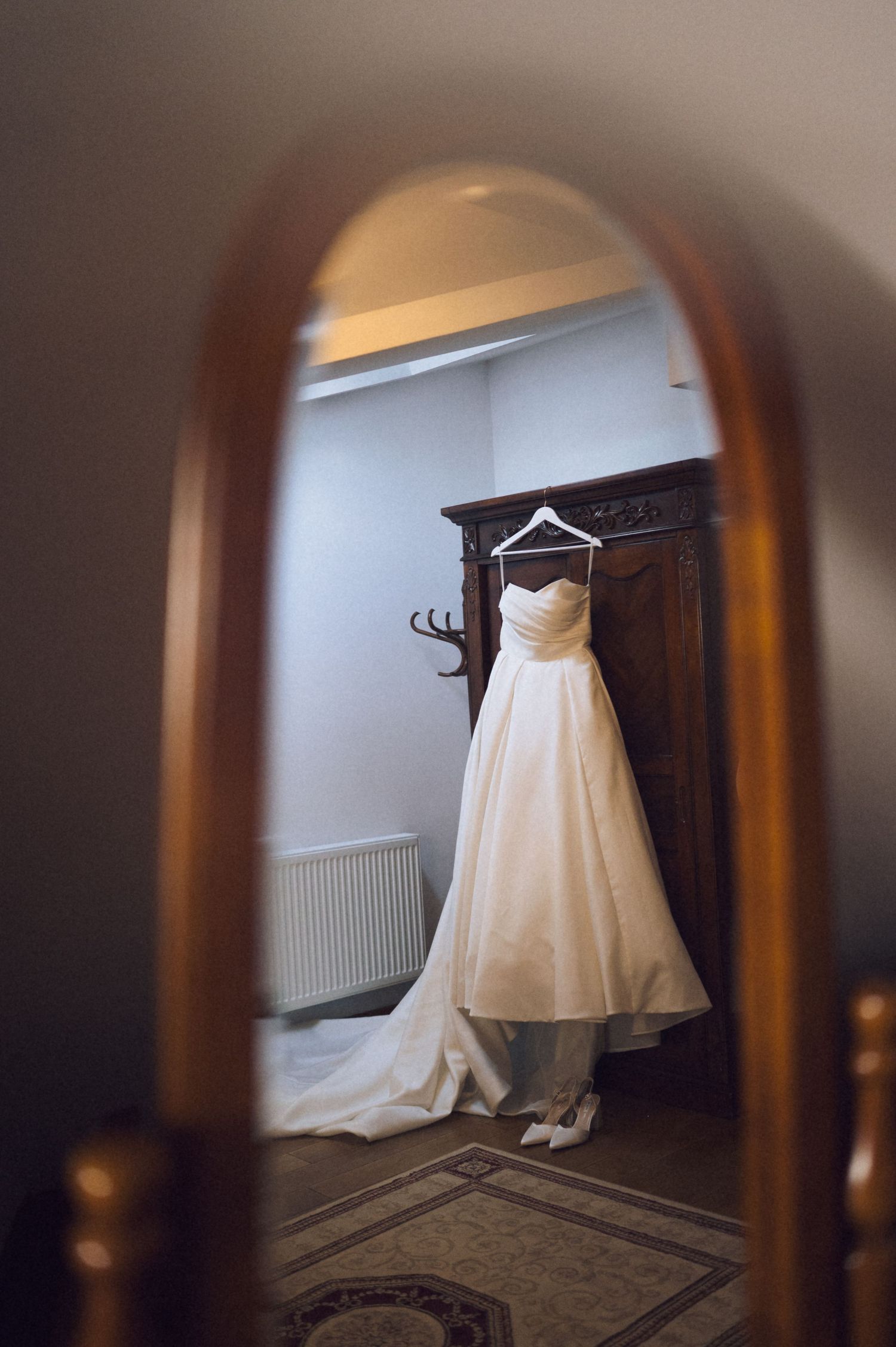 A white wedding dress hanging on a wooden wardrobe, reflected in an ornate wooden mirror frame.