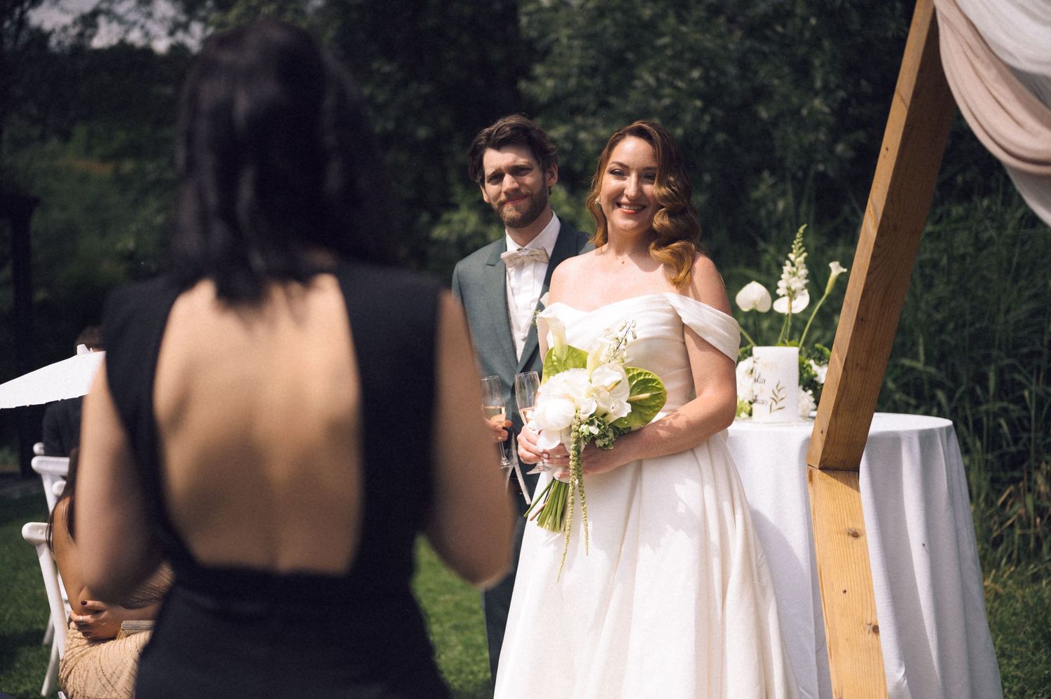 Bride in white gown holding bouquet smiles at groom during outdoor wedding ceremony with wooden arch backdrop.