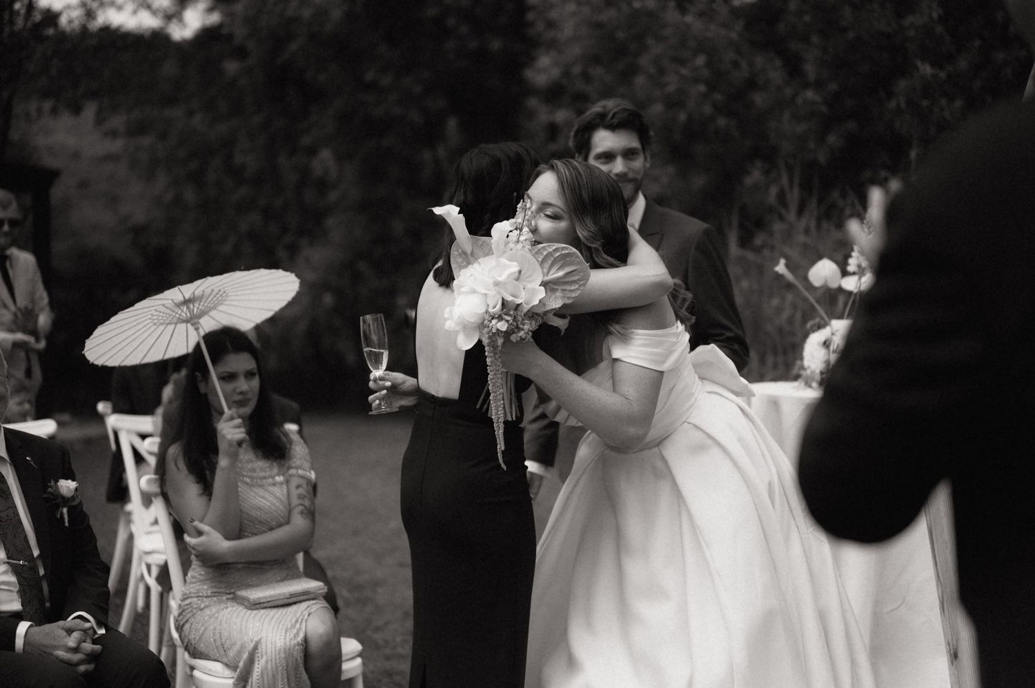 Black and white photo of a bride in a white gown embracing a guest holding a parasol at an outdoor wedding ceremony.