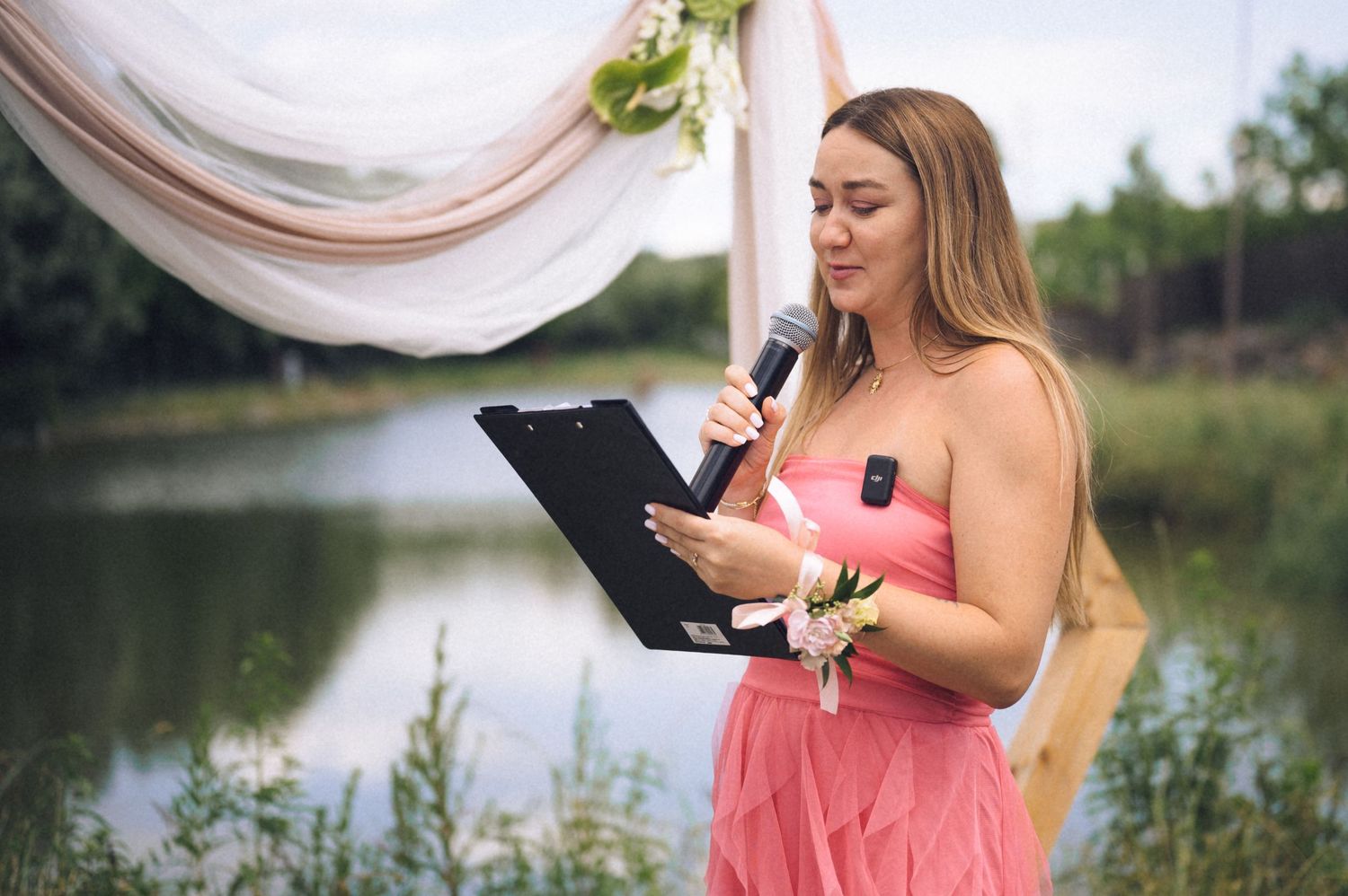 Woman in pink dress reads from clipboard at outdoor wedding ceremony near lake with floral arch backdrop.