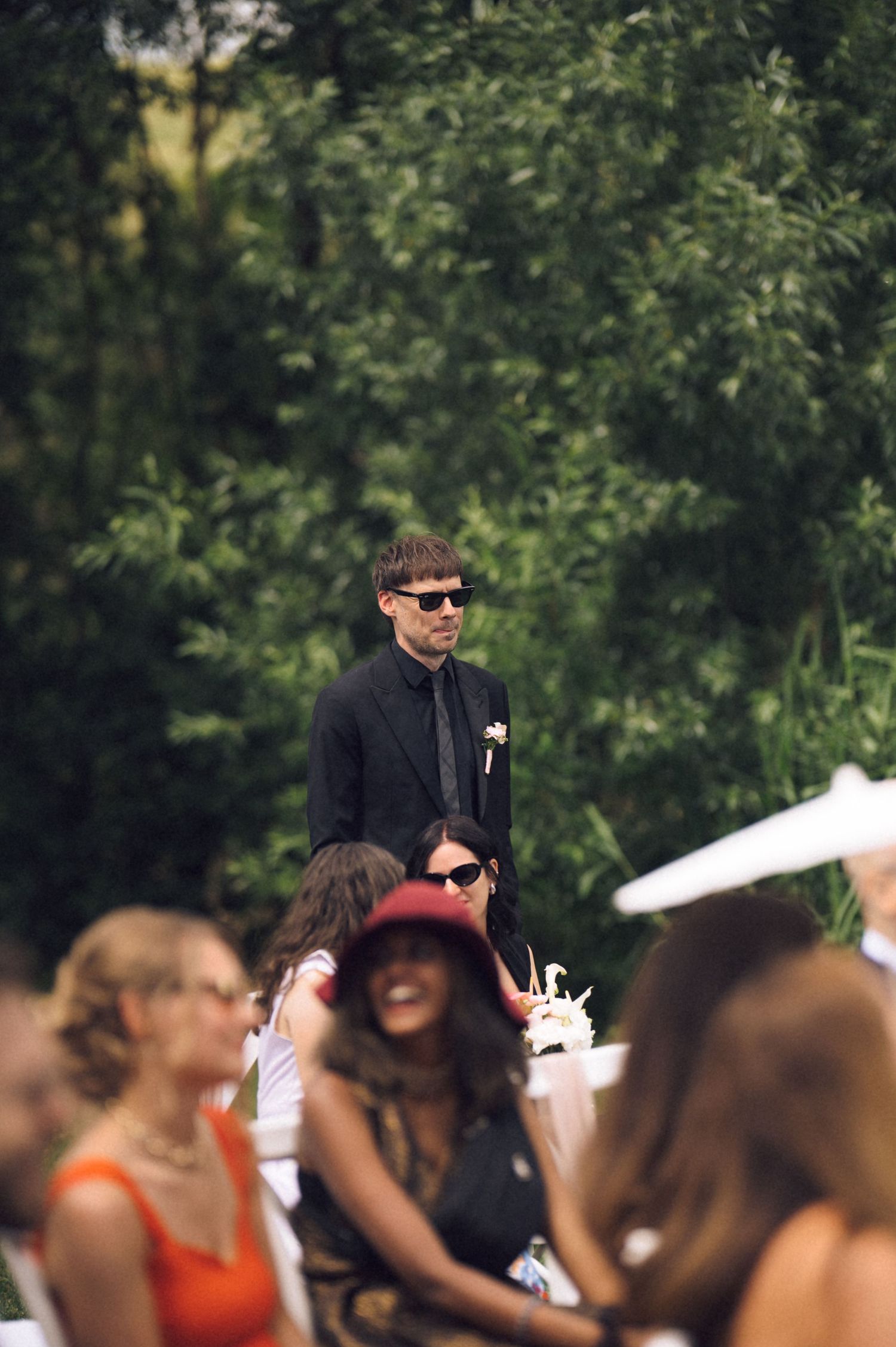 Man in black suit and sunglasses stands at outdoor wedding ceremony surrounded by seated guests and lush greenery.