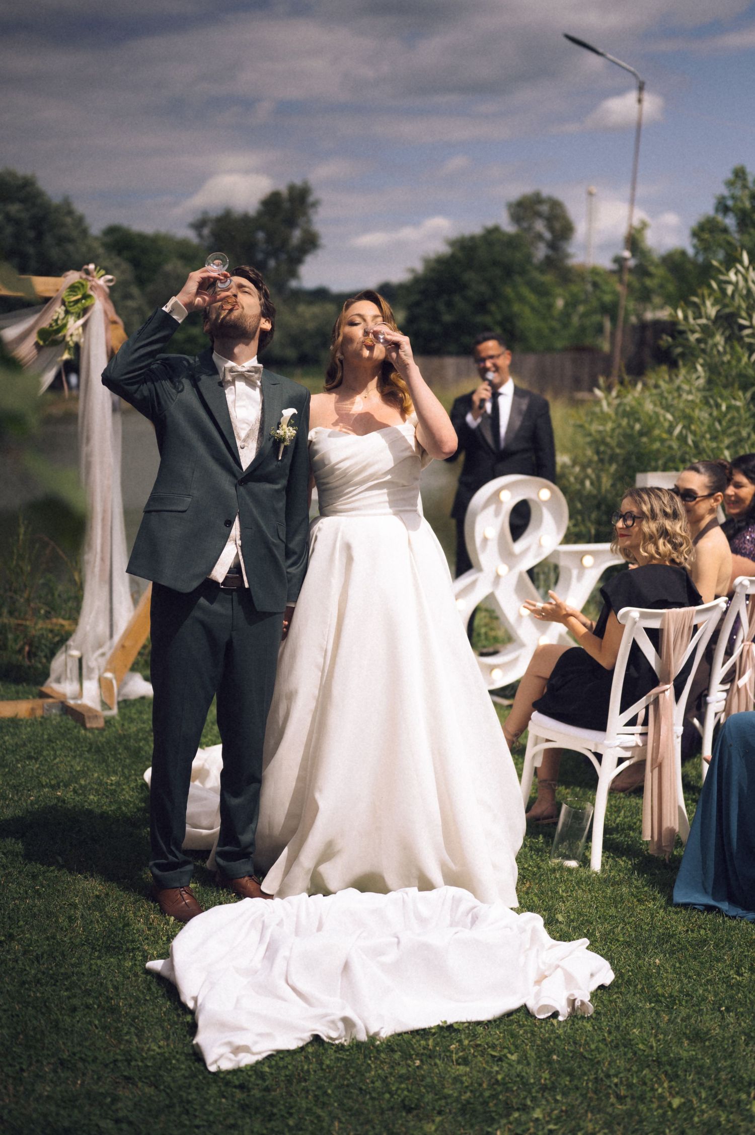 Bride and groom toasting with champagne glasses at outdoor wedding ceremony with guests seated nearby.