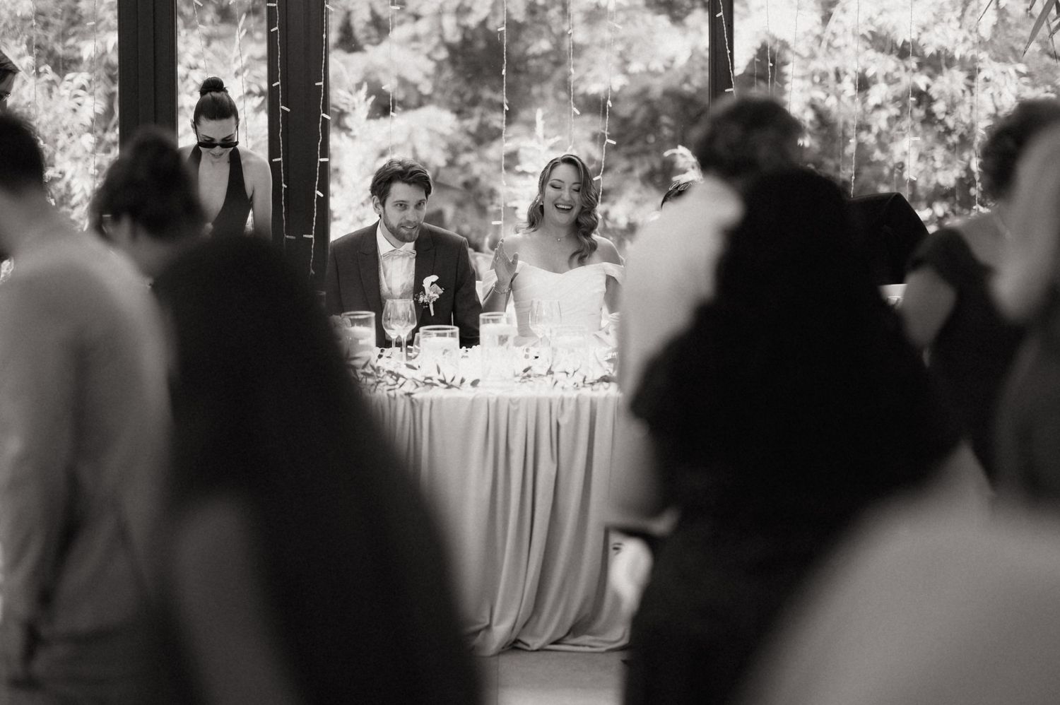 Black and white photo of a bride and groom smiling at their wedding reception sweetheart table surrounded by guests.