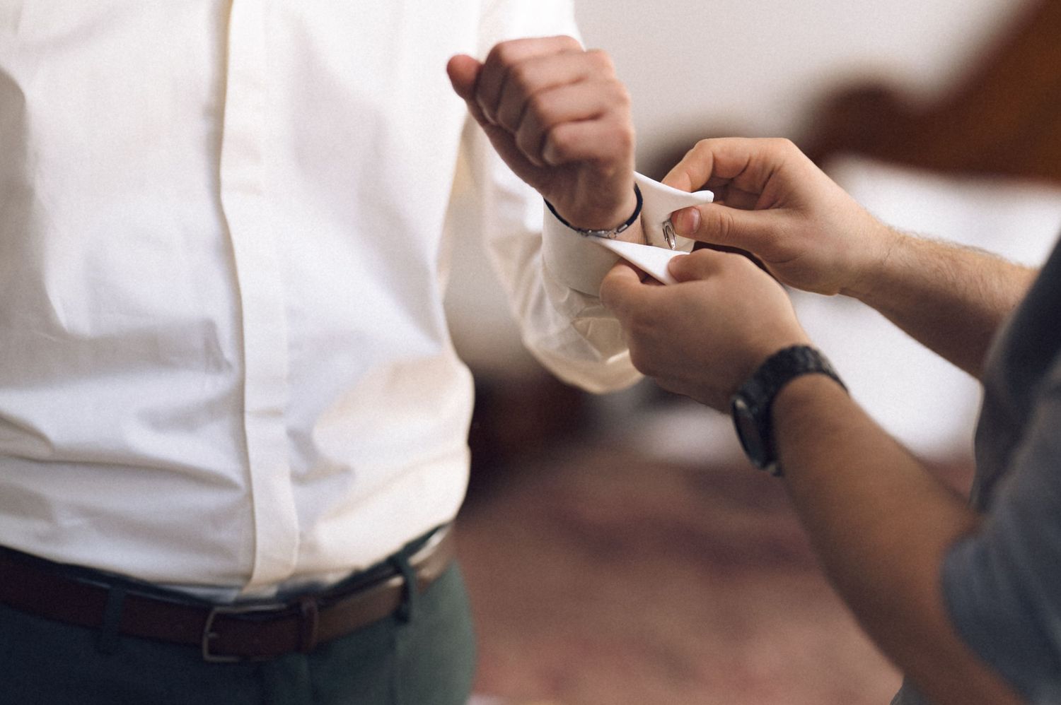 A person helping another put on cufflinks while dressed in a white dress shirt and brown belt.