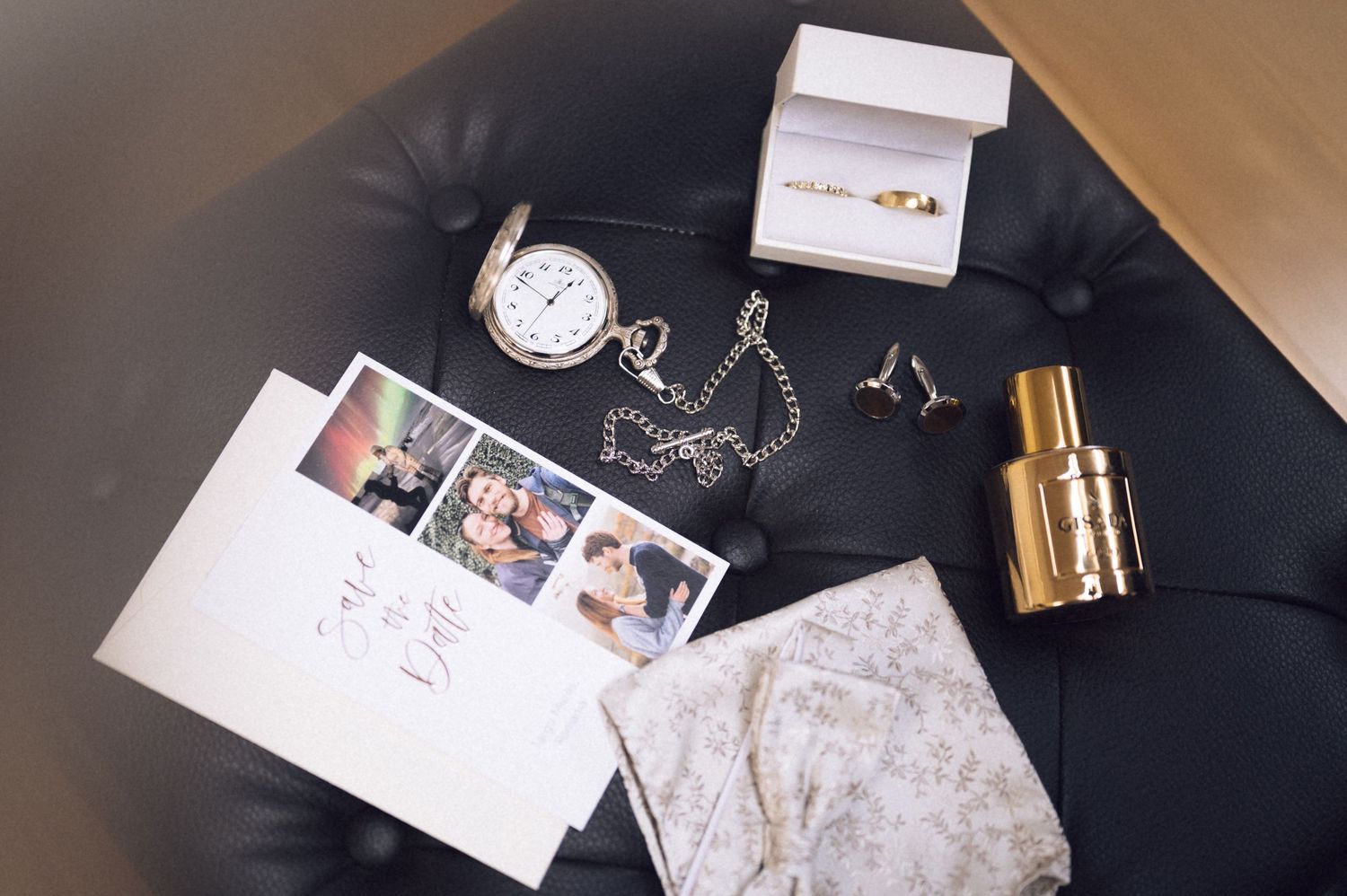 Flat lay of wedding accessories including pocket watch, rings, cufflinks, photos, and cologne on black leather surface.